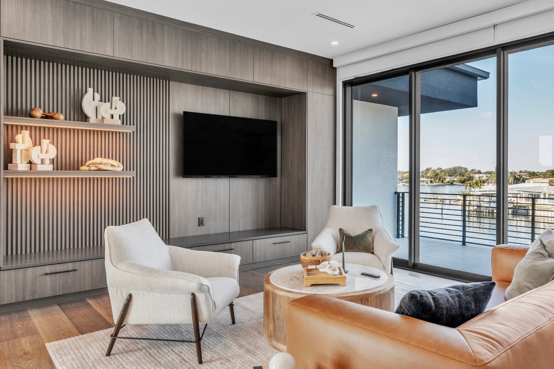 Waterfront living room with a gray built-in media wall, white lounge chairs, caramel leather sofa, and waterfront sliding doors